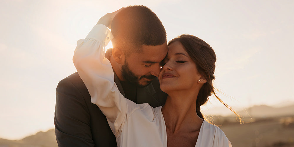 Casal, homem e mulher, abraçados de maneira afetuosa. Ambos são brancos. Ele veste uma roupa social preta e ela uma camisa branca